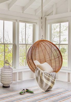 A wicker swing chair is hanging in the corner of a sunroom. There are pillows and blankets placed inside it. It is essential sunroom decor. 