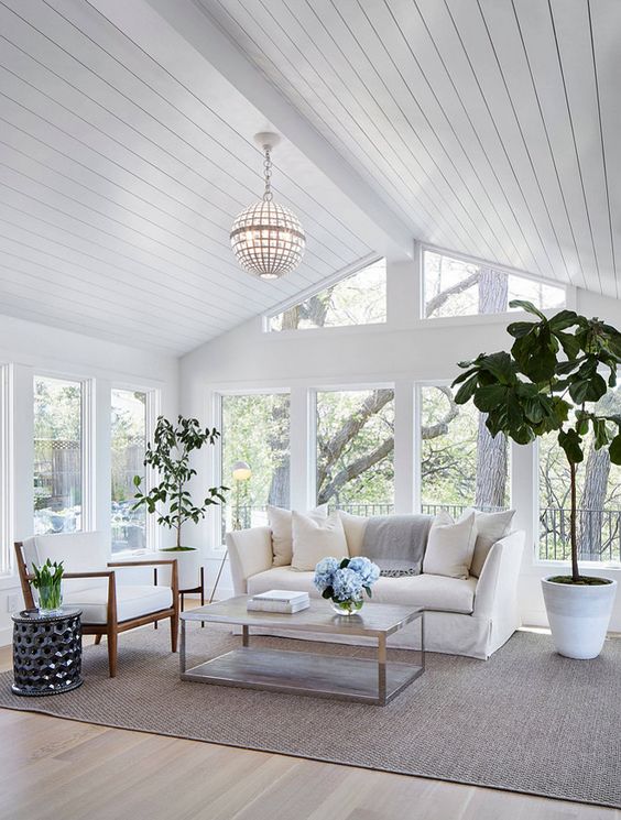 Plants are essential sunroom decor. In this photo, the interior of the sunroom is decorated with a sofa, coffee table, side chair and a tall planter tree sits next to the sofa.