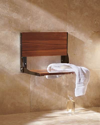 Folding Teak Wood Shower Seat attached to a shower wall. There is a white towel and soap set on the seat of the chair. 