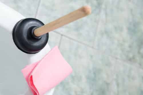 Ways to Clear Bathroom Clogs: Pictures is a black plunger with a wooden stick handle next to a pink cleaning rag. 