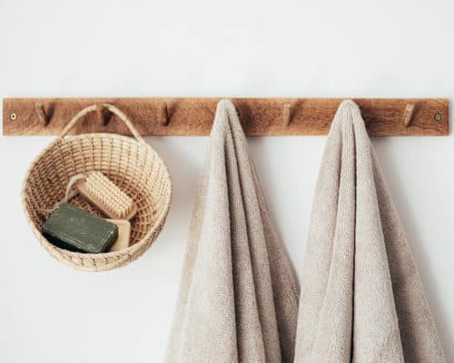 Items Your Shouldn't Store in Your Bathroom: Two towels and a basket are hanging from a horizontal, wooden towel rack. There is a bar of soap and scrub brush inside the hanging basket. 