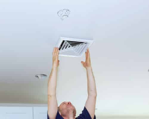 A man is reaching up to place the vent cover on a bathroom exhaust fan. The fan is in the ceiling.