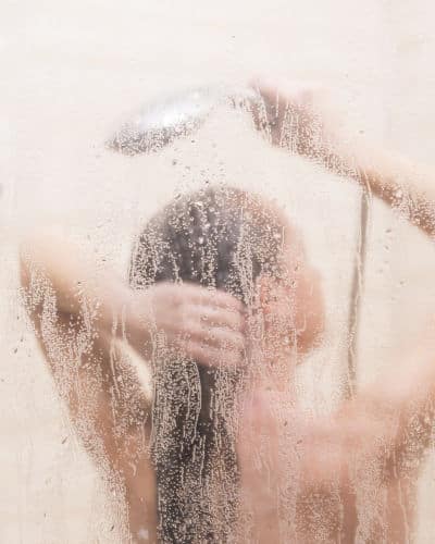 Bathroom Exhaust Fan: Shower glass is dripping with condensation. Behind the glass is a woman, rinsing her hair. One hand is on her hair while the other is holding a shower head above it. 